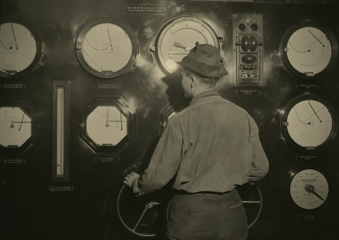 Archival photograph, 1942: a man in a hat standing at a control panel covered in large round and octagonal instrument dials, his back to the camera, one hand on a valve wheel.