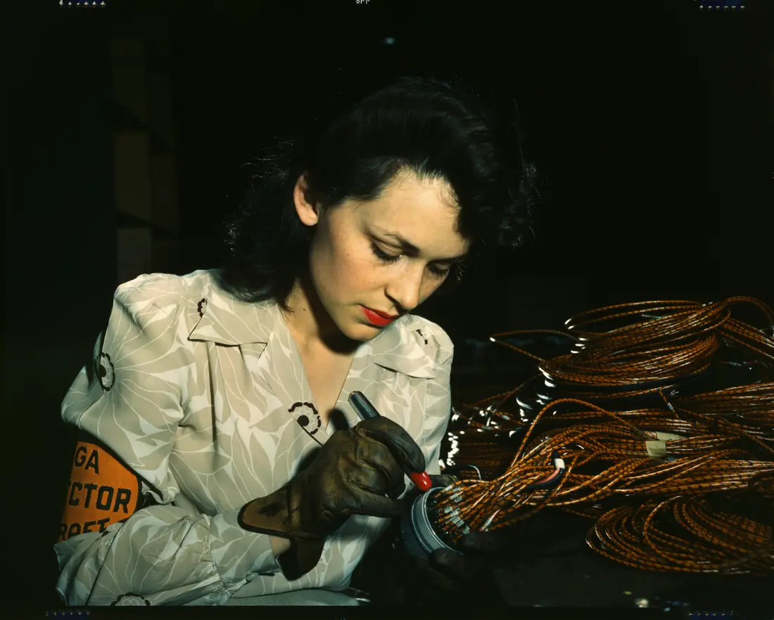 Archival colour photograph, 1942: a factory worker in a patterned shirt bent over a bundle of copper-coloured wiring, working it with a hand tool under a single light in a near-black room.