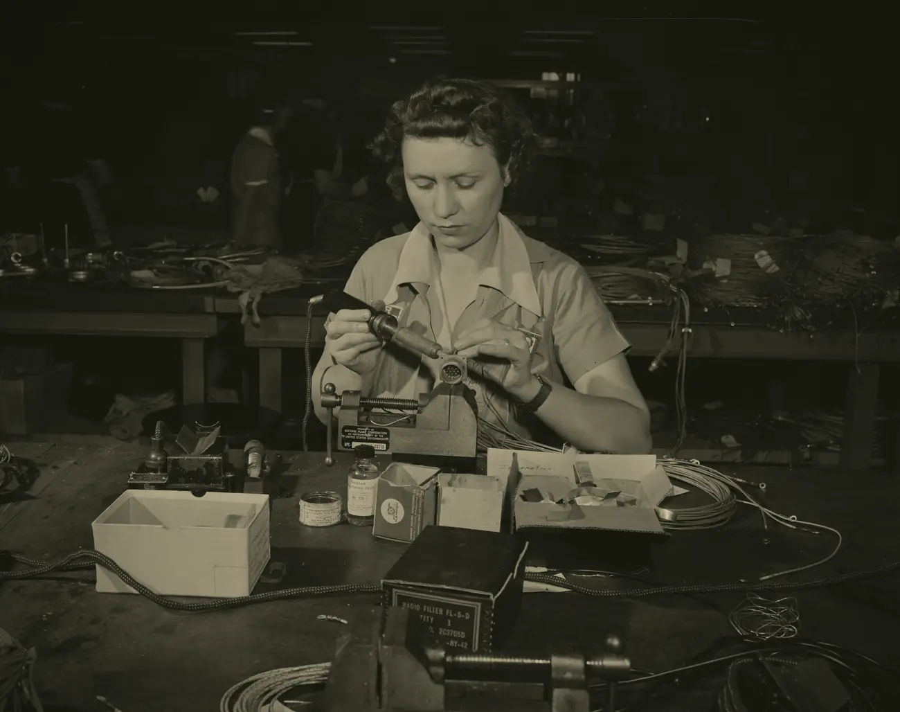 Archival photograph, 1942: a woman at a long factory bench soldering a contact onto the end of a wire, a lit soldering iron in one hand, spools of cable and small parts spread across the bench in front of her.